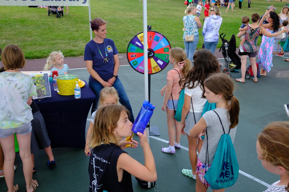 a female in a Be Well Northborough shirt looks at a prize wheel while surrounded by children
