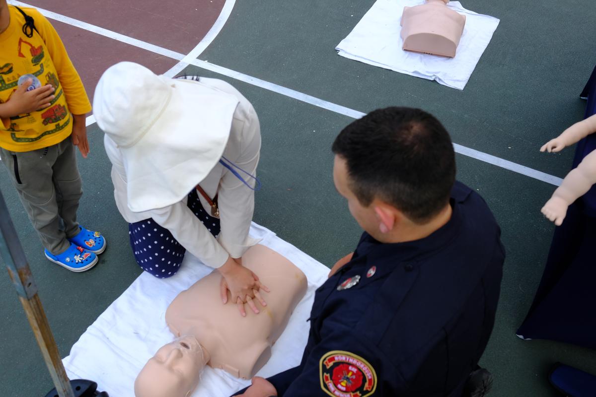 A child kneeling in front of a CPR dummy while a fireman in uniform looks on.