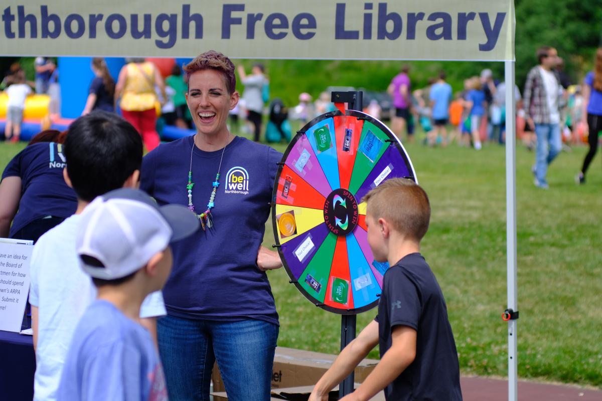 Female in a Be Well Northborough shirt laughs with children next to a prize wheel in front of a library banner